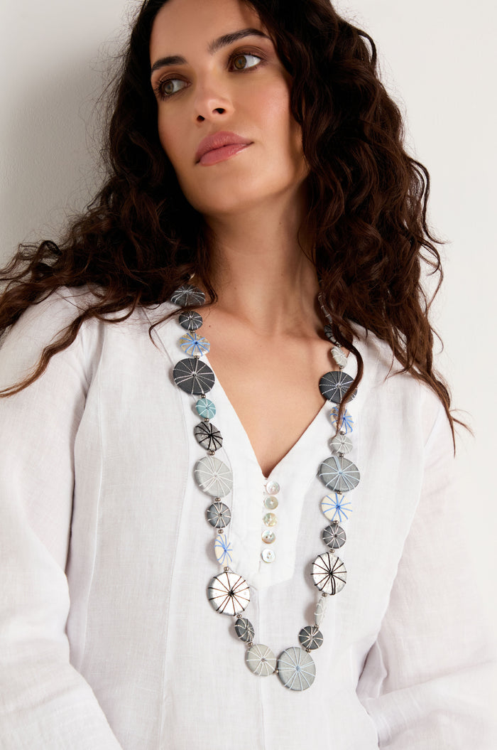 A woman with long curly hair, dressed in a white blouse, stands against a white wall wearing the Long Wooden Metallic Parasols Necklace.