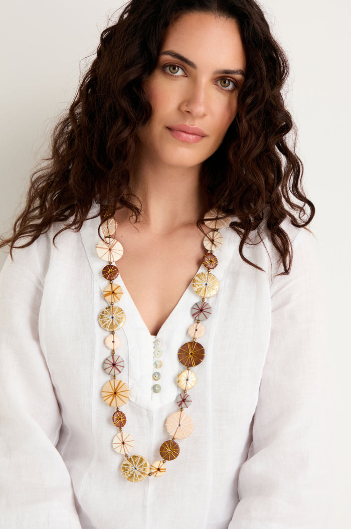 Woman with long curly brown hair, wearing a white blouse with cotton detail and a Long Wooden Metallic Parasols Necklace, stands against a plain light background.