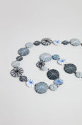 The Long Wooden Metallic Parasols Necklace features round, flat beads in various sizes with geometric line patterns in blue, gray, black, and white, arranged spirally on a plain background.