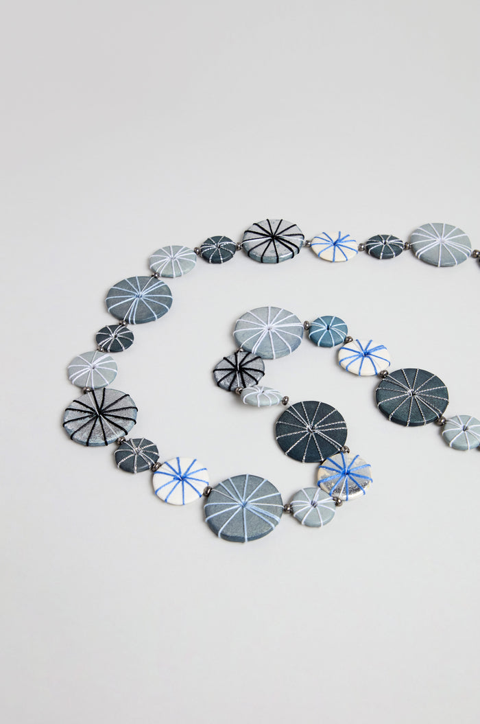 The Long Wooden Metallic Parasols Necklace features round, flat beads in various sizes with geometric line patterns in blue, gray, black, and white, arranged spirally on a plain background.