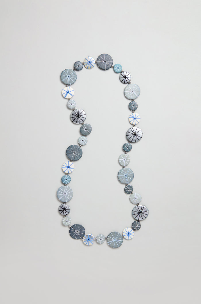 The Long Wooden Metallic Parasols Necklace features round, flat beads in gray, blue, and white with line patterns and subtle cotton accents, arranged in a loose oval on a plain background.