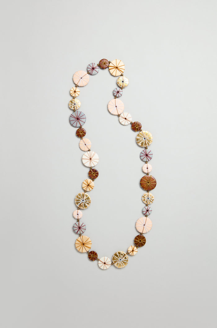 The Long Wooden Metallic Parasols Necklace is a long necklace with round, flat beads in pastel pink, beige, gold, and brown, arranged irregularly on a light gray cotton-accented background.