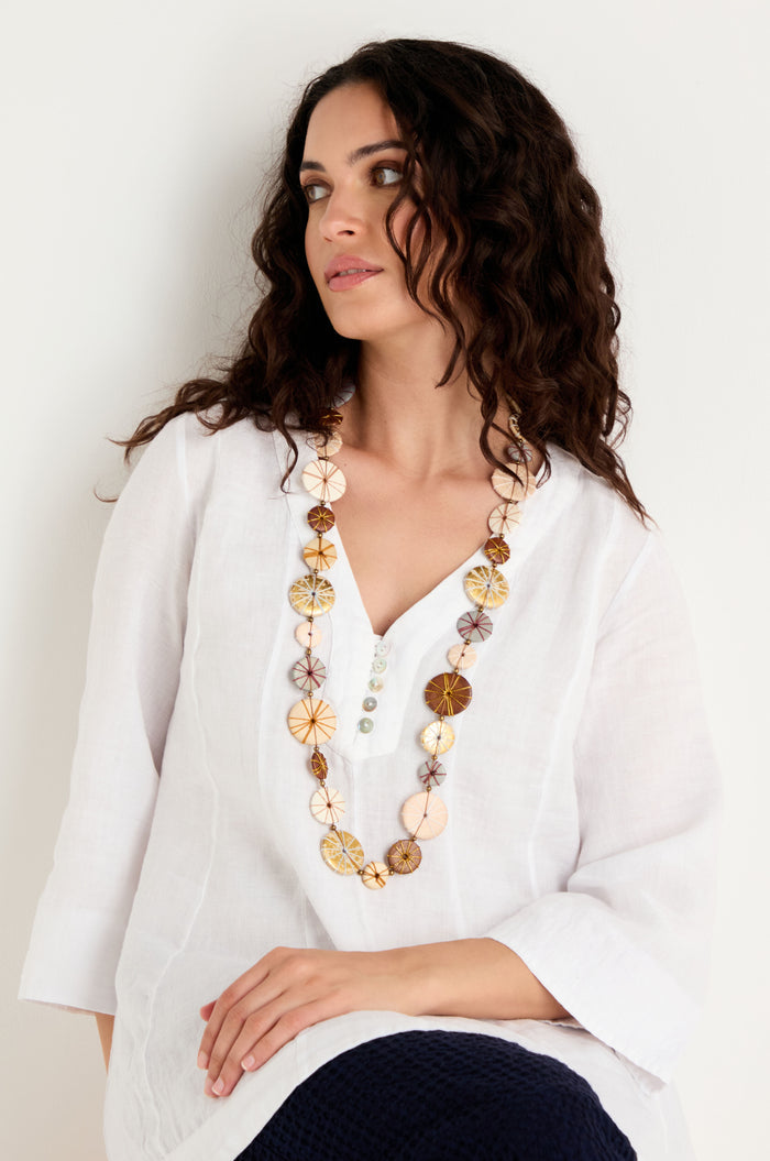 A woman with long curly hair wears a white tunic and the Long Wooden Metallic Parasols Necklace, featuring round decorative beads, while sitting against a plain white background.