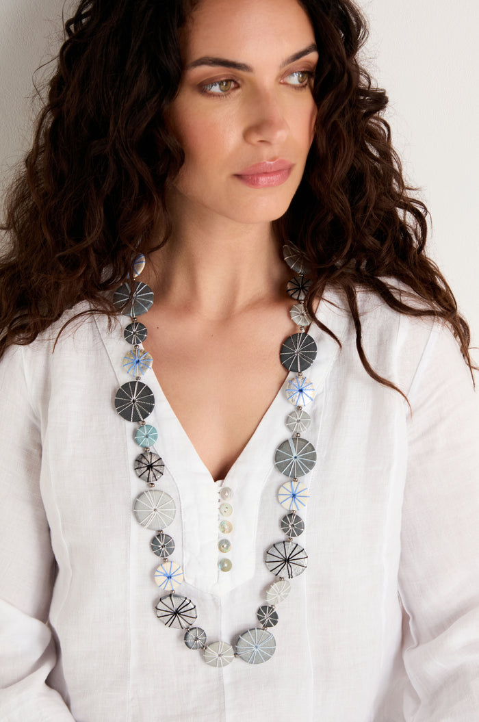 A woman with long curly hair wears a white blouse and the Long Wooden Metallic Parasols Necklace, which features black, white, and blue patterned beads. She is looking slightly to the side.