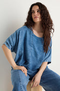A woman with long curly hair sits on a wooden stool against a plain light background, wearing the Lyocell Panelled Boxy Top and matching denim pants with panelled details.