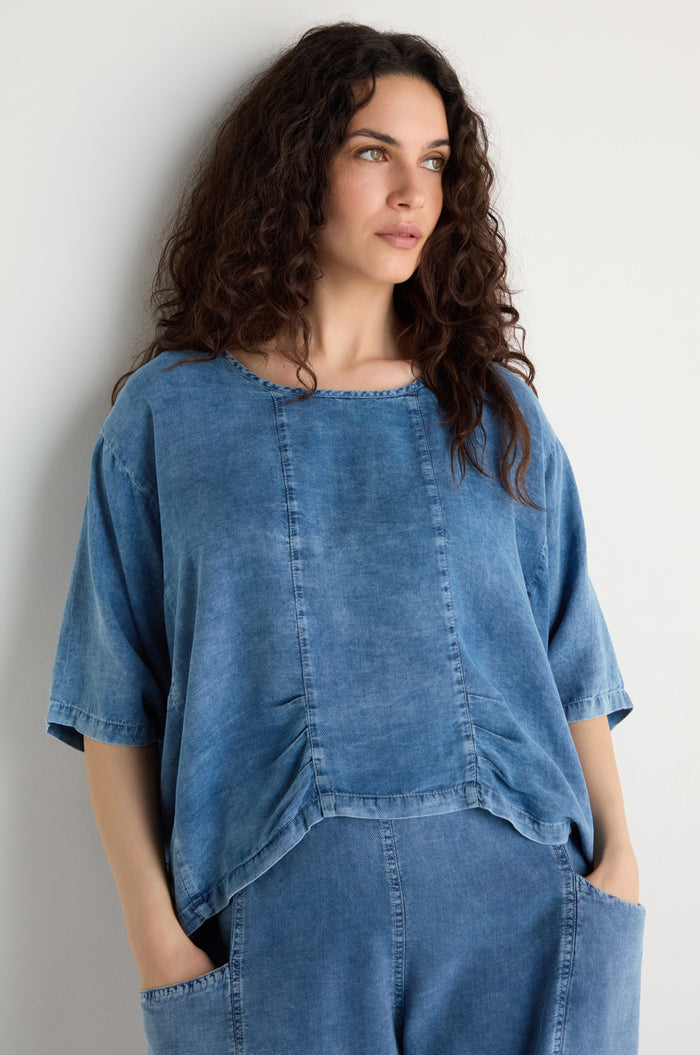 A woman with long curly hair, hands in her pockets, stands against a plain white wall wearing the Lyocell Panelled Boxy Top and matching pants.
