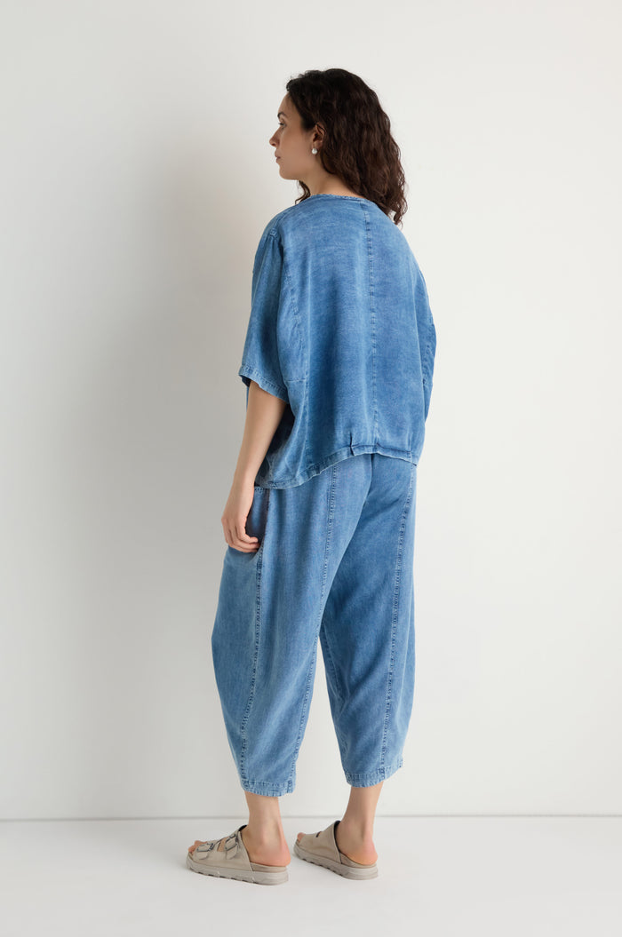 A person in a loose-fitting blue Lyocell Panelled Boxy Top and matching denim pants stands facing a white wall, their back turned slightly to the camera.