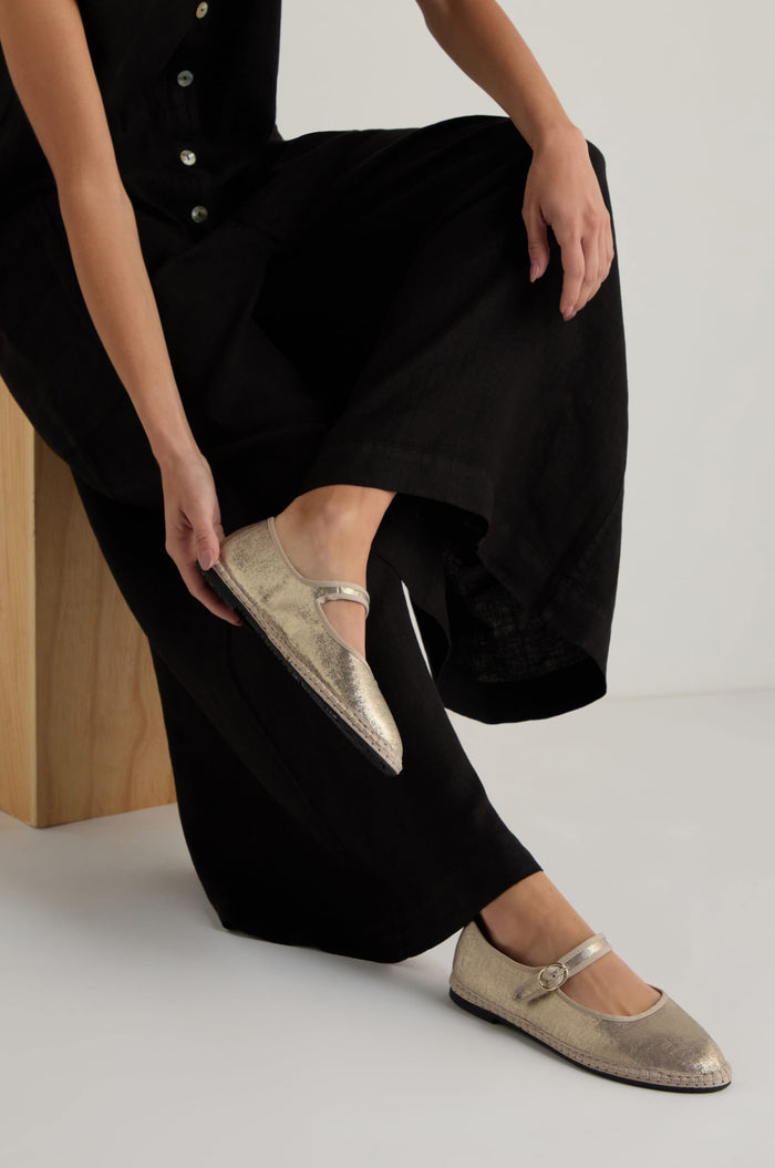 Someone in black pants sits on a wooden bench, adjusting their Metallic Mary Jane Shoes, which feature a recycled bicycle tyre sole.