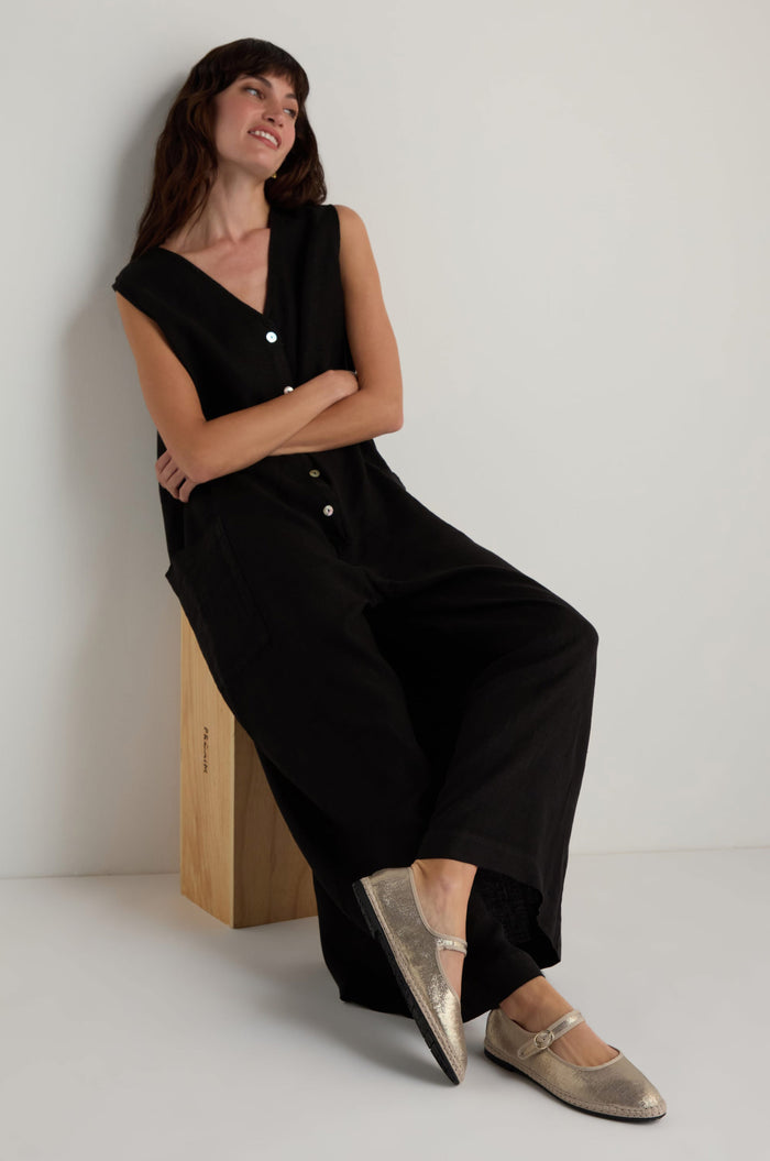 A woman with arms crossed sits on a wooden block against a white wall, wearing a sleeveless black jumpsuit and Metallic Mary Jane Shoes. Her legs are casually extended.