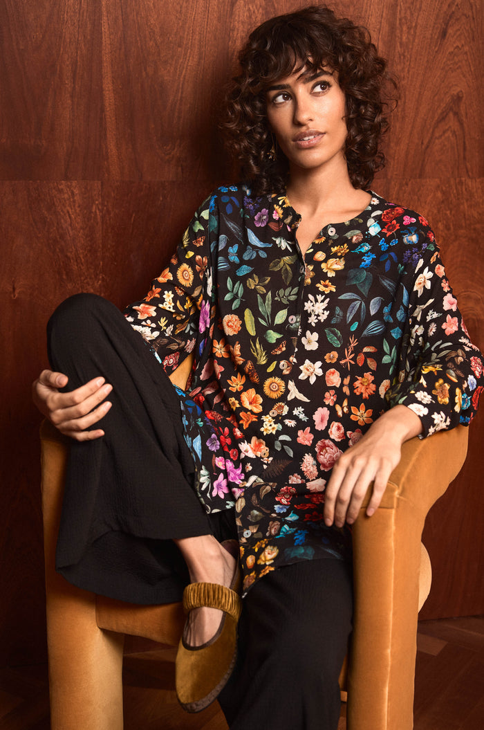 A person with curly hair sits in a tan armchair, wearing the Midnight Garden Viscose Shirt, black pants, and brown flat shoes, against a wooden wall.