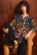 A person with curly hair sits in a tan armchair, wearing the Midnight Garden Viscose Shirt, black pants, and brown flat shoes, against a wooden wall.