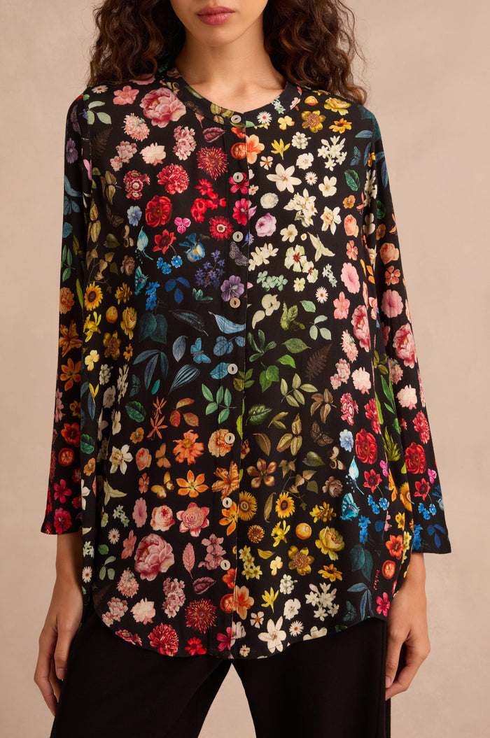 A woman stands against a neutral background, wearing the Midnight Garden Viscose Shirt—a black long-sleeve blouse featuring a colorful floral print and a subtly flared silhouette.