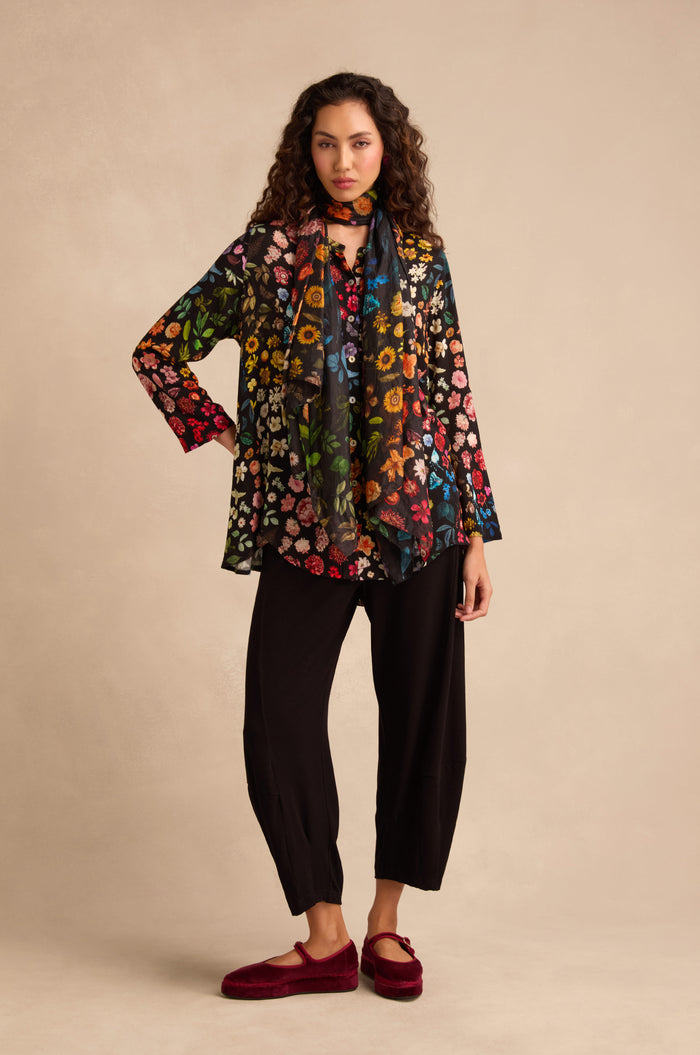A woman stands against a beige background, dressed in the Midnight Garden Viscose Shirt with a flared silhouette, paired with a matching floral scarf, loose black pants, and red slippers.