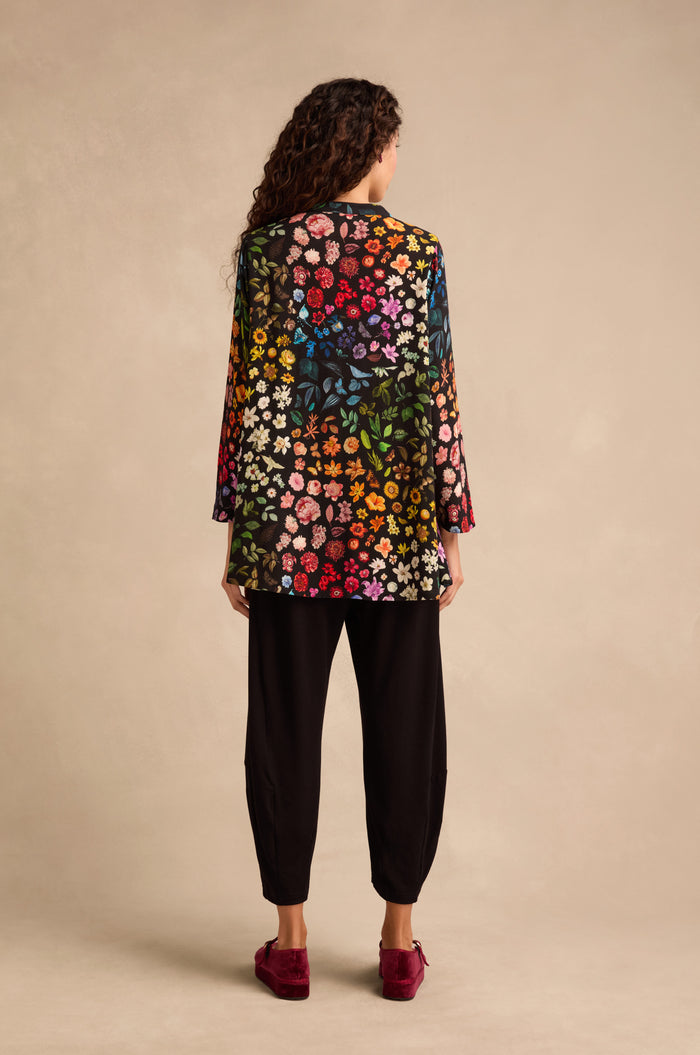 A person stands facing away, showcasing the Midnight Garden Viscose Shirt—known for its vibrant floral pattern and flared silhouette—paired with black pants and red shoes. Their curly hair is visible from behind, set against a beige background.