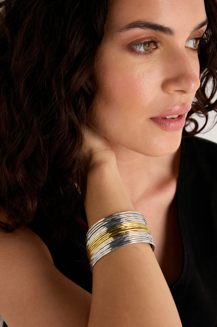 A woman with wavy dark hair, dressed in a black top, rests her hand on her neck while wearing the Mixed Metal Cuff.