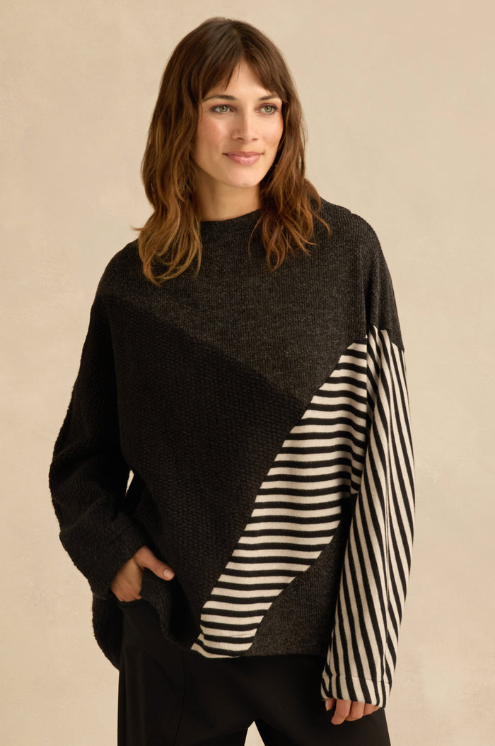 A woman is wearing the Mixed Stripe Top—an oversized black sweater with asymmetric panel construction and diagonal black and white striped panels on one sleeve and part of the front—standing with one hand in her pocket.
