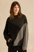 A woman is wearing the Mixed Stripe Top—an oversized black sweater with asymmetric panel construction and diagonal black and white striped panels on one sleeve and part of the front—standing with one hand in her pocket.