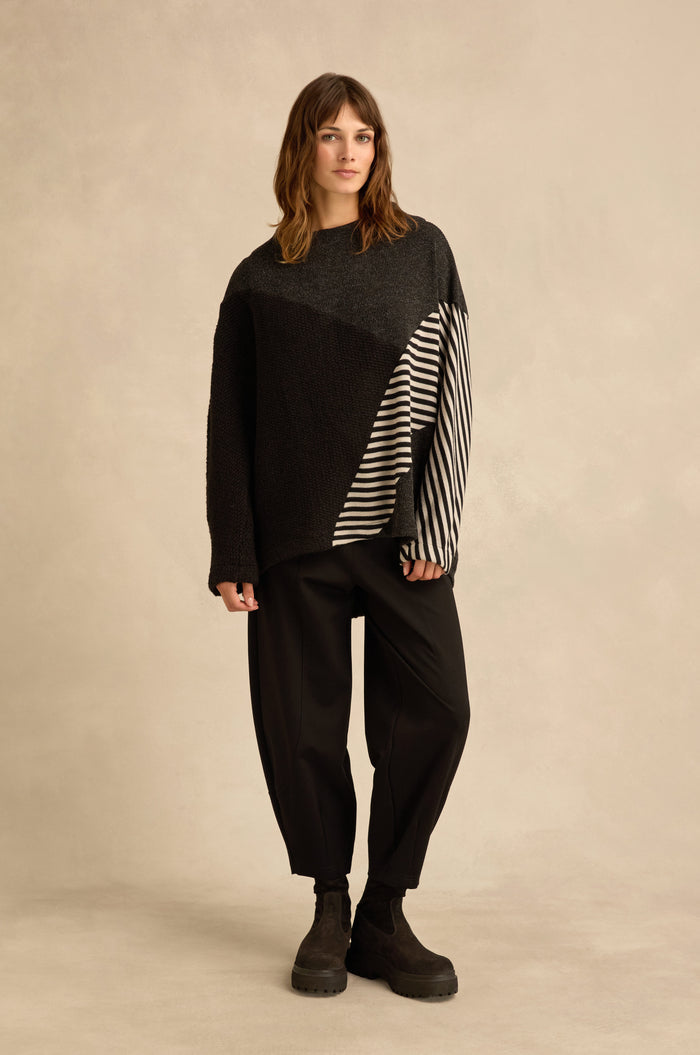 A woman stands against a beige background wearing the Mixed Stripe Top—an oversized black and gray sweater with a striped sleeve and asymmetric panel construction—paired with black pants and black boots.