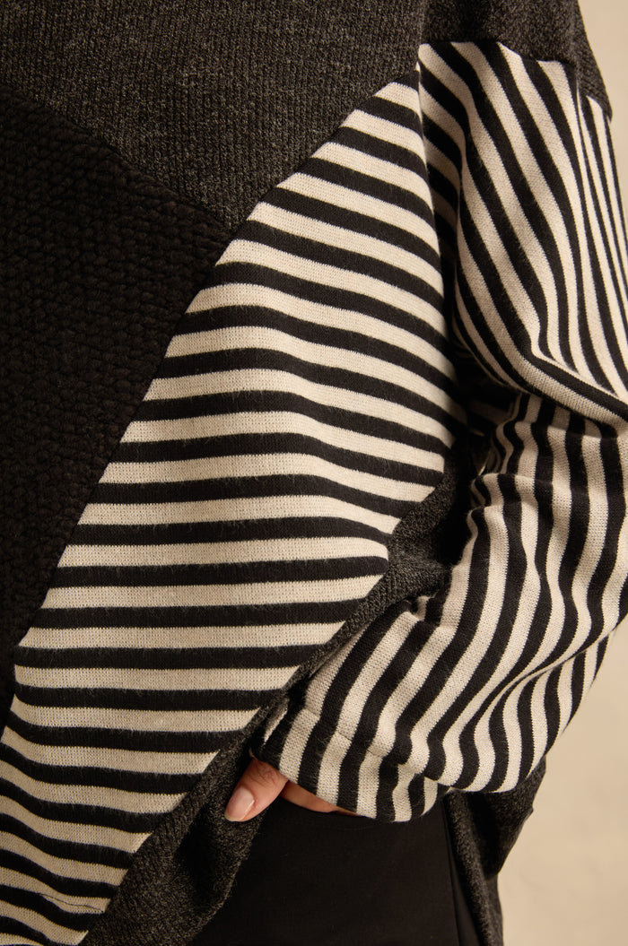A person wearing the Mixed Stripe Top, an oversized sweater with asymmetric panel construction that combines a solid black section and a black-and-white horizontal stripe section, with one hand partially visible near the waist.