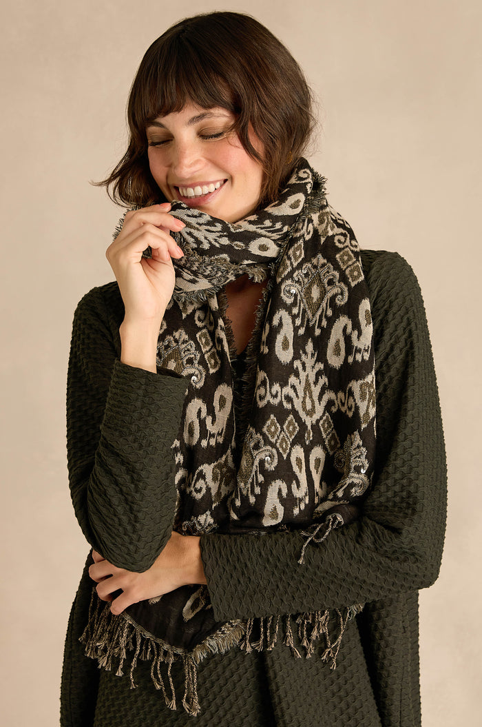 A woman smiles with her eyes closed, standing against a neutral palette background, wearing the Motif Sequin Scarf styled with a textured dark green cotton-wool blend sweater.