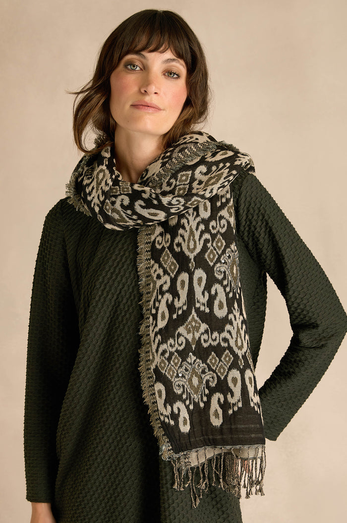 A woman stands against a neutral background wearing a textured dark green dress made from a cozy cotton-wool blend, styled with the Motif Sequin Scarf—a patterned black and white scarf featuring fringed ends.