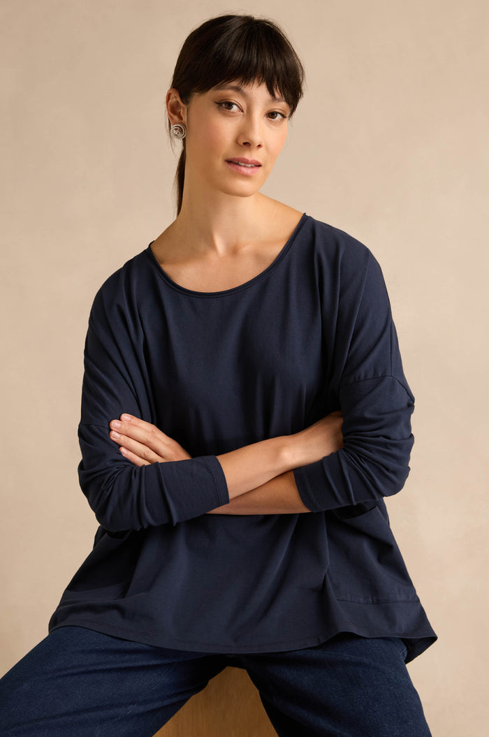 A woman with straight dark hair, wearing the Navy Cotton Jersey Pocket Top and jeans, sits with her arms crossed against a plain beige background.