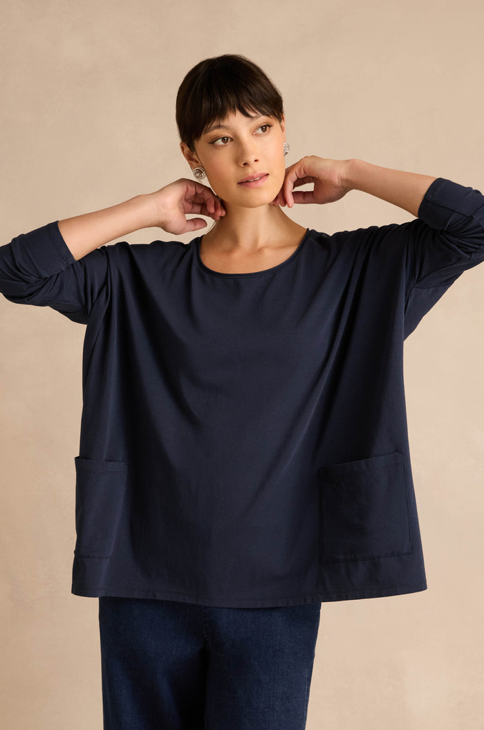 A woman stands against a beige background, wearing the Navy Cotton Jersey Pocket Top with front pockets and dark pants, her hands held near her neck.