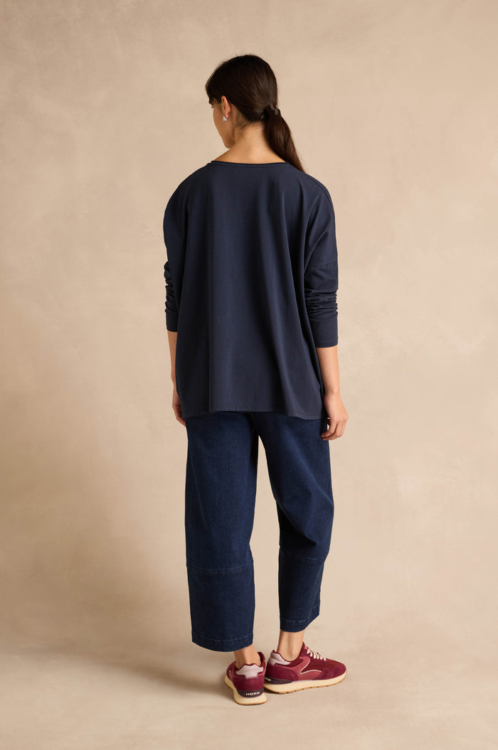A person with long brown hair in a ponytail wears the Navy Cotton Jersey Pocket Top styled oversized, along with wide-leg denim pants and burgundy sneakers, standing with their back to the camera against a beige background.