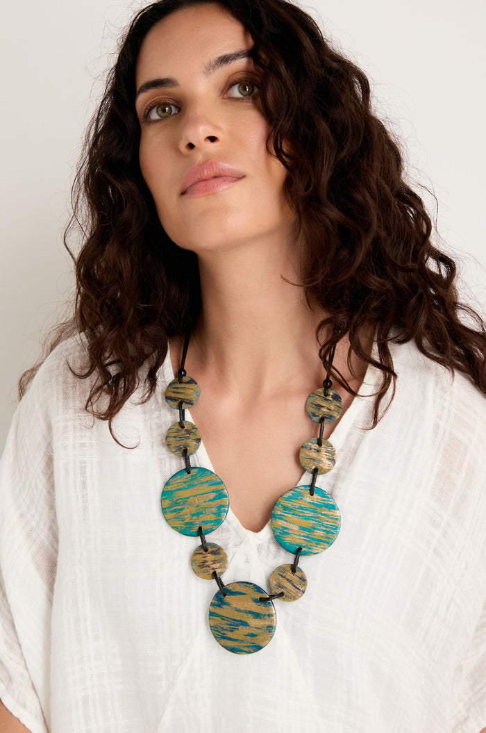 A woman with wavy brown hair wears a white textured top and the Painted Disc Necklace, featuring large round and oval blue and gold beads.