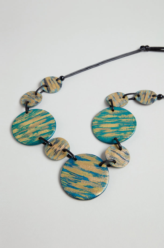 The Painted Disc Necklace features round, flat teal beads with gold brushstroke patterns, strung on a black cord and displayed against a light background.