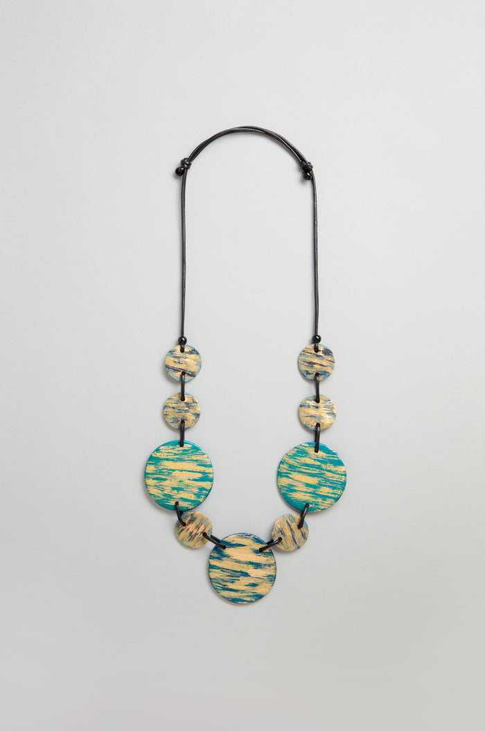 The Painted Disc Necklace features seven round, flat beads in blue and beige patterns on a black adjustable cord, shown against a plain light background.