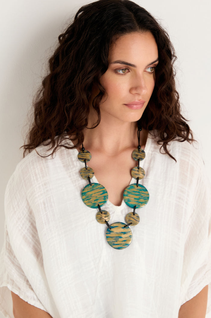 Woman with wavy dark hair wears a white textured blouse and the Painted Disc Necklace, featuring large blue and green circular accents, standing against a plain white background.