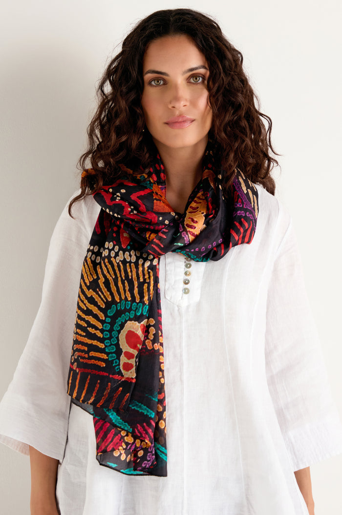 A woman styled in a white tunic wears the Painted Firework Silk Scarf, featuring an abstract print, while standing against a plain light-colored background.