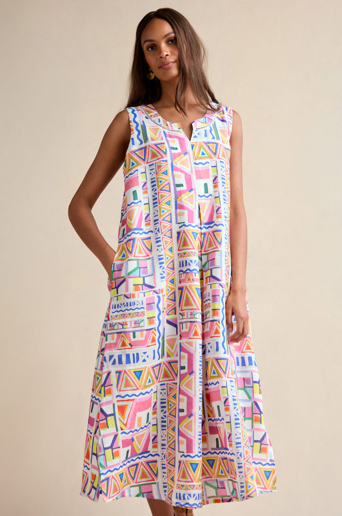 A woman models the Painted Geo Linen Dress, a sleeveless and loose-fitting linen piece featuring a vibrant geometric print, set against a plain beige background.