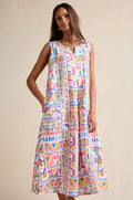 A woman models the Painted Geo Linen Dress, a sleeveless and loose-fitting linen piece featuring a vibrant geometric print, set against a plain beige background.
