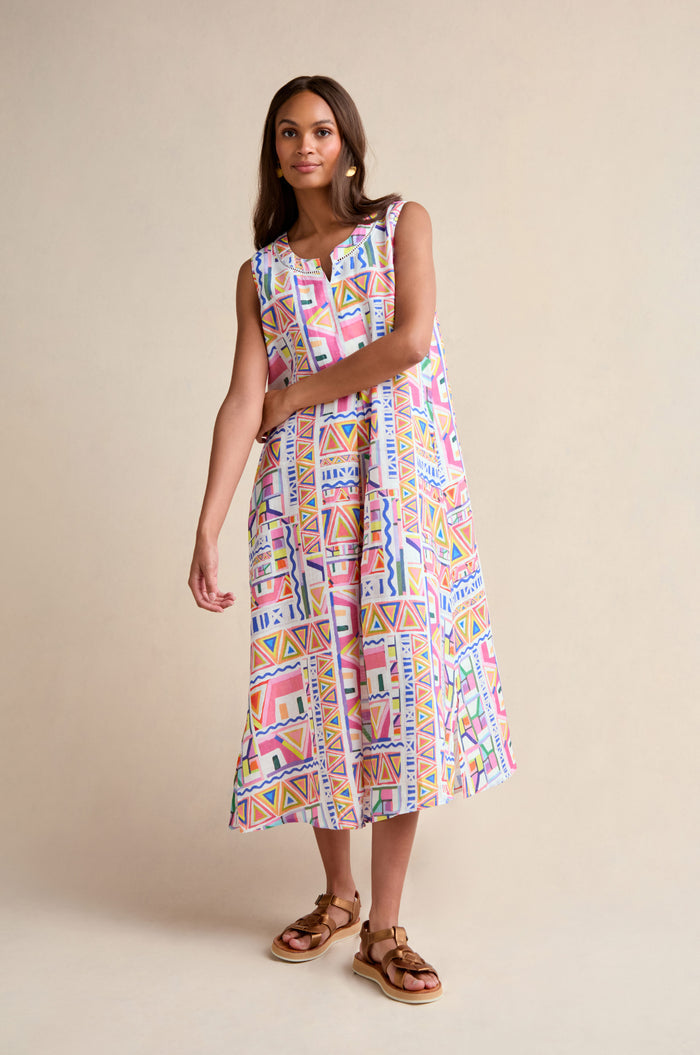 A woman wears the Painted Geo Linen Dress, a sleeveless linen piece featuring a vibrant geometric print, paired with brown sandals and set against a beige background.