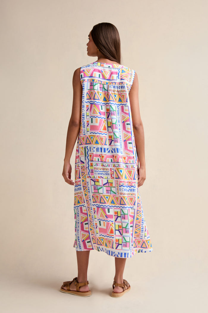 A woman faces away, wearing the Painted Geo Linen Dress—a sleeveless, midi-length linen dress with a colorful geometric print—paired with brown sandals against a plain light background.