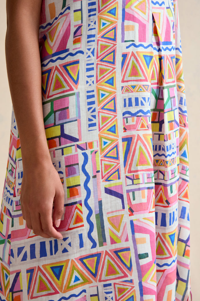 A person wears the Painted Geo Linen Dress, showcasing a vibrant geometric print with triangles, rectangles, and wavy lines in red, pink, green, blue, and yellow. Only the torso and arm are visible.