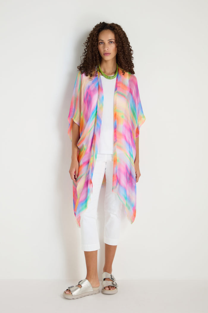 A woman stands against a white background wearing the Painted Horizon Kimono—a colorful, patterned open front layer—over a white top and cropped pants, paired with sandals and layered necklaces.