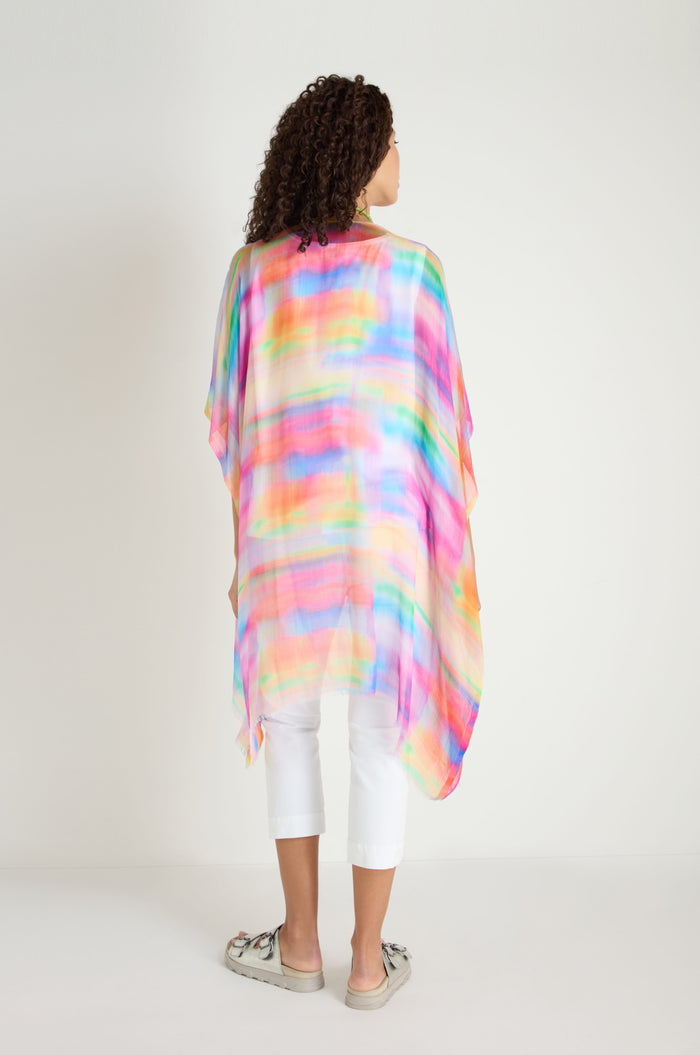 A woman stands against a white background, shown from behind, wearing the colorful Painted Horizon Kimono with white cropped pants and light-colored shoes.