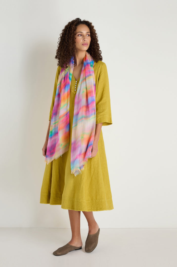 A woman in a loose yellow dress and brown shoes stands against a plain wall, wearing the Painted Horizon Scarf—a vibrant, lightweight accessory.