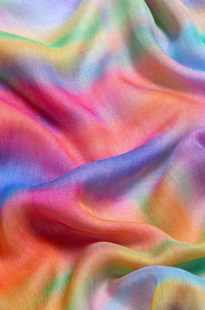 The Painted Horizon Scarf features a smooth, wavy tie-dye fabric in blended pink, orange, blue, green, and purple shades. Crafted from luxurious modal and silk, this lightweight scarf adds vibrant color to any outfit.