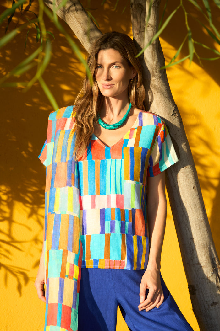 A woman stands in sunlight by a yellow wall and tree, wearing a colorful top, blue pants, green necklace, and the Painted Stripes Scarf draped elegantly around her shoulders.
