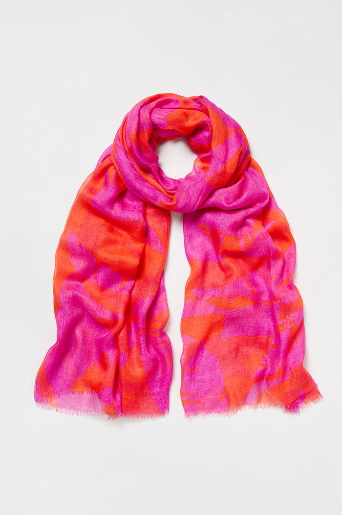The Palm Mirage Scarf is a lightweight modal silk blend featuring an abstract tropical foliage pattern in vibrant pink and orange, shown loosely folded on a plain white background.