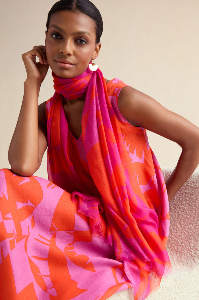 A woman wearing a sleeveless pink and orange patterned dress with the Palm Mirage Scarf sits on a textured chair, looking at the camera.