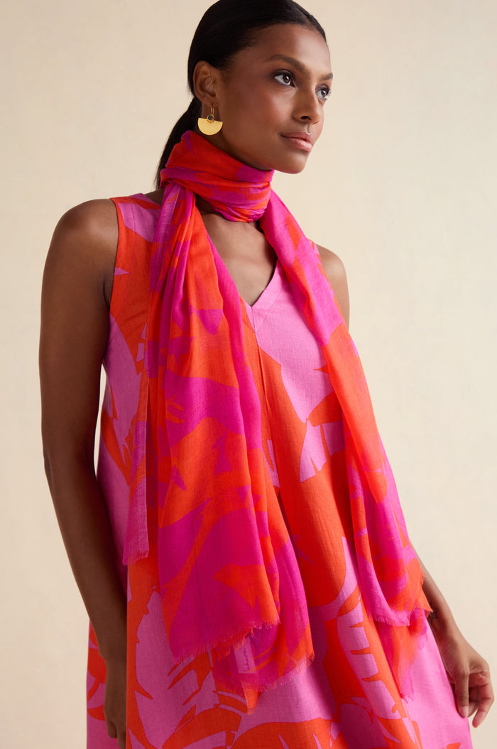 A woman with straight dark hair and gold earrings wears a sleeveless pink and orange dress, accessorized with the Palm Mirage Scarf in matching pink and coral shades. She has a neutral expression.