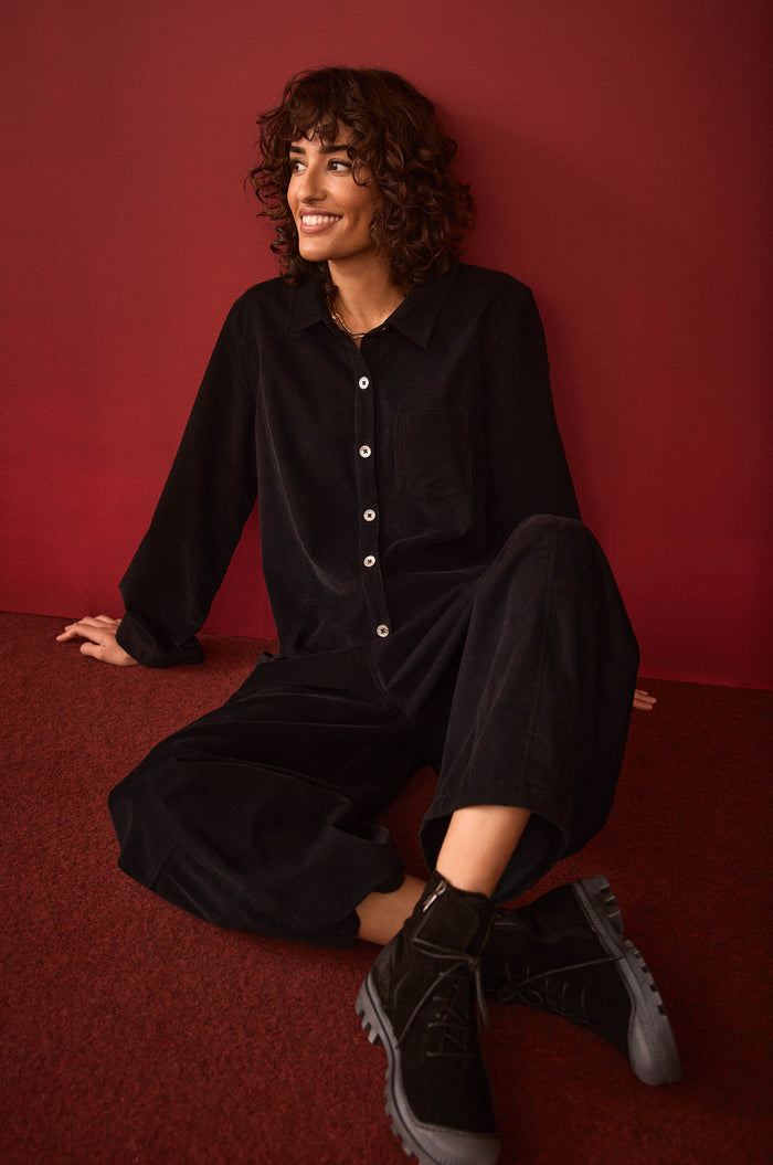 A person with curly hair sits on a red floor against a red wall, smiling and looking slightly to the side while wearing the Pinwale Cord Jumpsuit and boots.