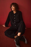 A person with curly hair sits on a red floor against a red wall, smiling and looking slightly to the side while wearing the Pinwale Cord Jumpsuit and boots.