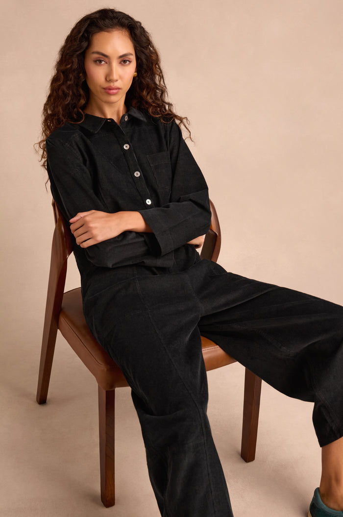 A person with long curly hair sits on a wooden chair, wearing the Pinwale Cord Jumpsuit—black, pure cotton, with bubble leg detail—arms crossed and a neutral expression, against a beige background.