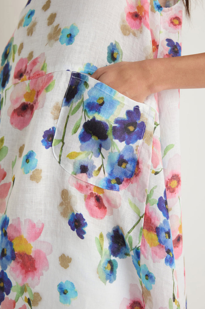 Someone slips a hand into the side pocket of a Primrose Print Sleeveless Linen Tunic, featuring white linen with vibrant pink, blue, and purple primrose patterns.